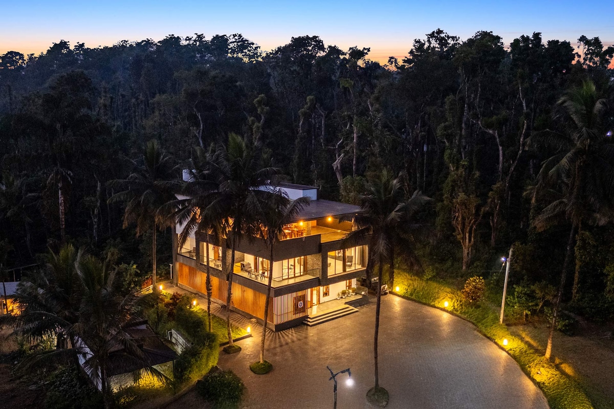 An aerial view captures the villa's modern architecture amidst tall trees as the sky transitions to twilight. Soft lighting enhances the building's exterior, while landscaped gardens frame the entrance, creating an inviting atmosphere in the serene hill surroundings.