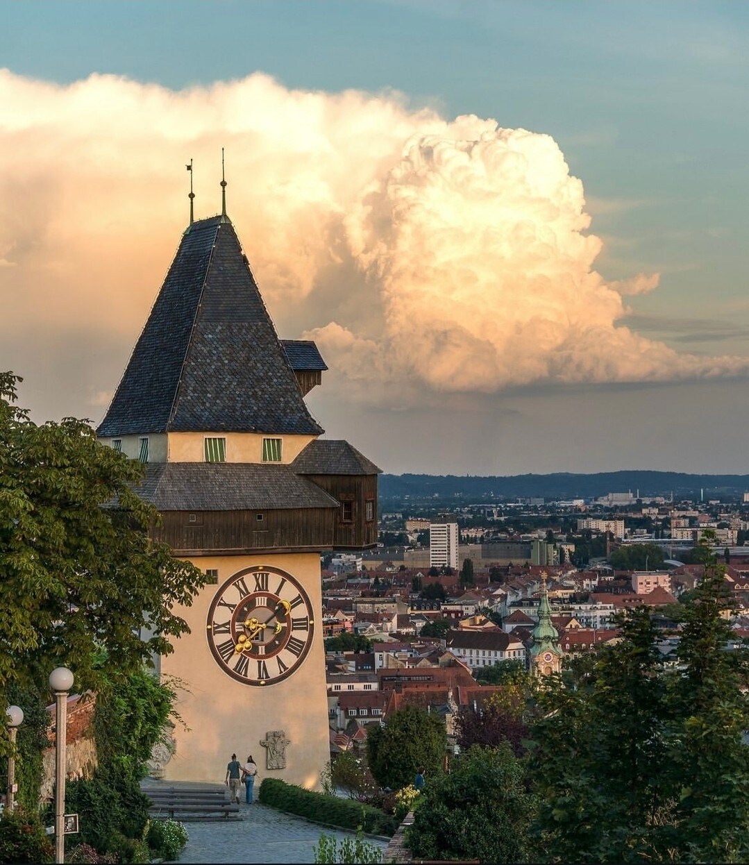 The iconic clock tower stands prominently, its distinct architecture highlighted against the sky. Lush greenery surrounds the base, while expansive views of the city and distant clouds provide a dramatic backdrop, showcasing the blend of nature and urban landscape.