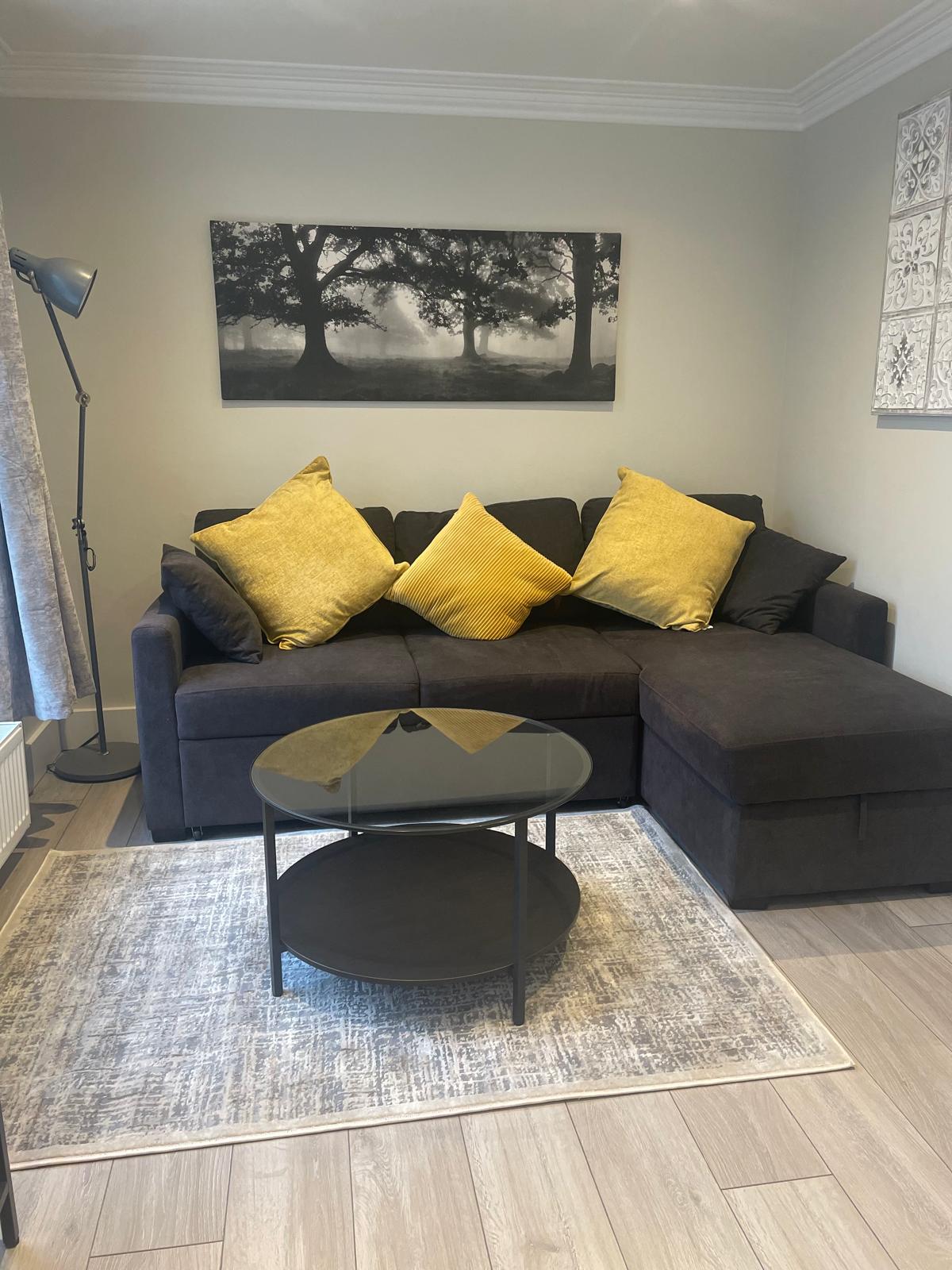 A comfortable sitting area is presented with a dark sofa adorned with yellow cushions. A round, black coffee table is positioned on a soft area rug, while a floor lamp stands to one side, providing additional lighting. A black-and-white artwork of trees is displayed on the wall.