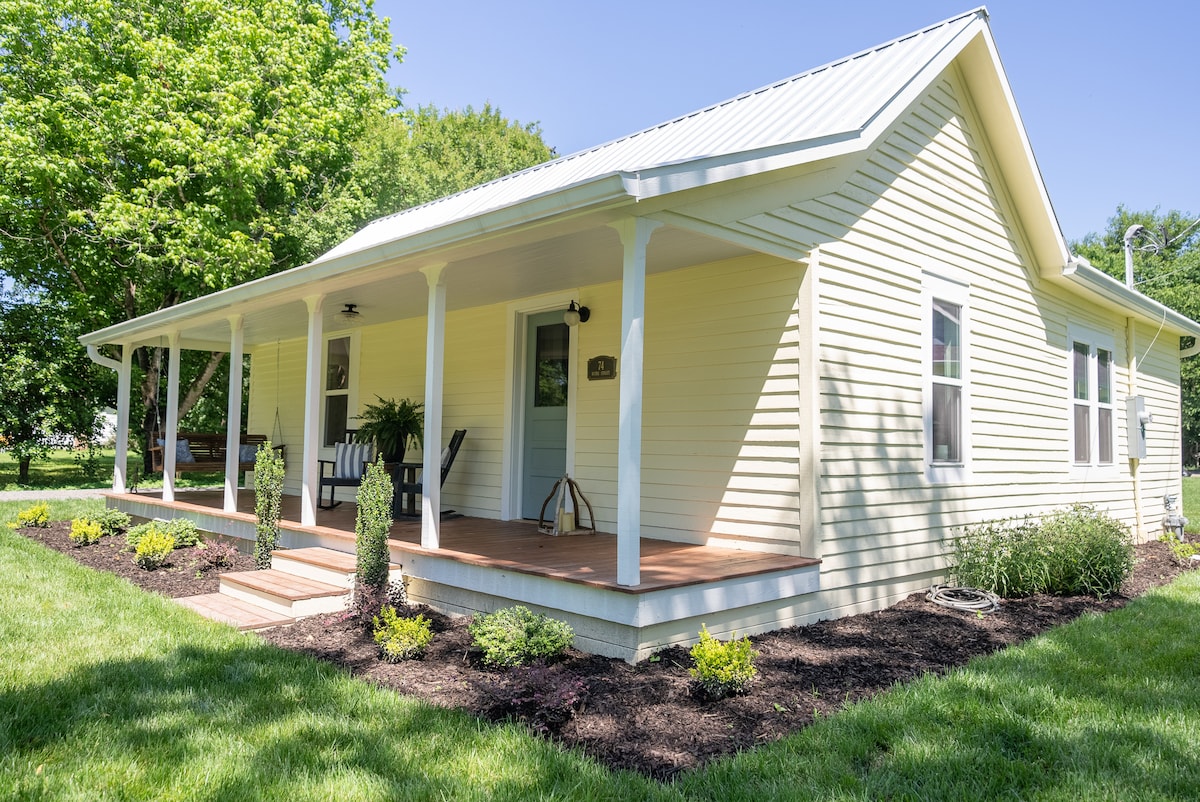 The exterior of a charming cottage is showcased with a welcoming porch featuring rocking chairs. A well-maintained lawn surrounds the house, complemented by neatly arranged shrubs and flower beds. The building has a light-colored facade and a sloping roof under a clear blue sky.