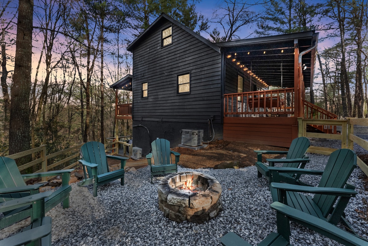 A cozy outdoor firepit area is surrounded by comfortable Adirondack chairs, set on a gravel surface. The cabin structure features dark siding and multiple windows, with a spacious deck visible in the background, all framed by trees as the evening sky transitions to twilight.