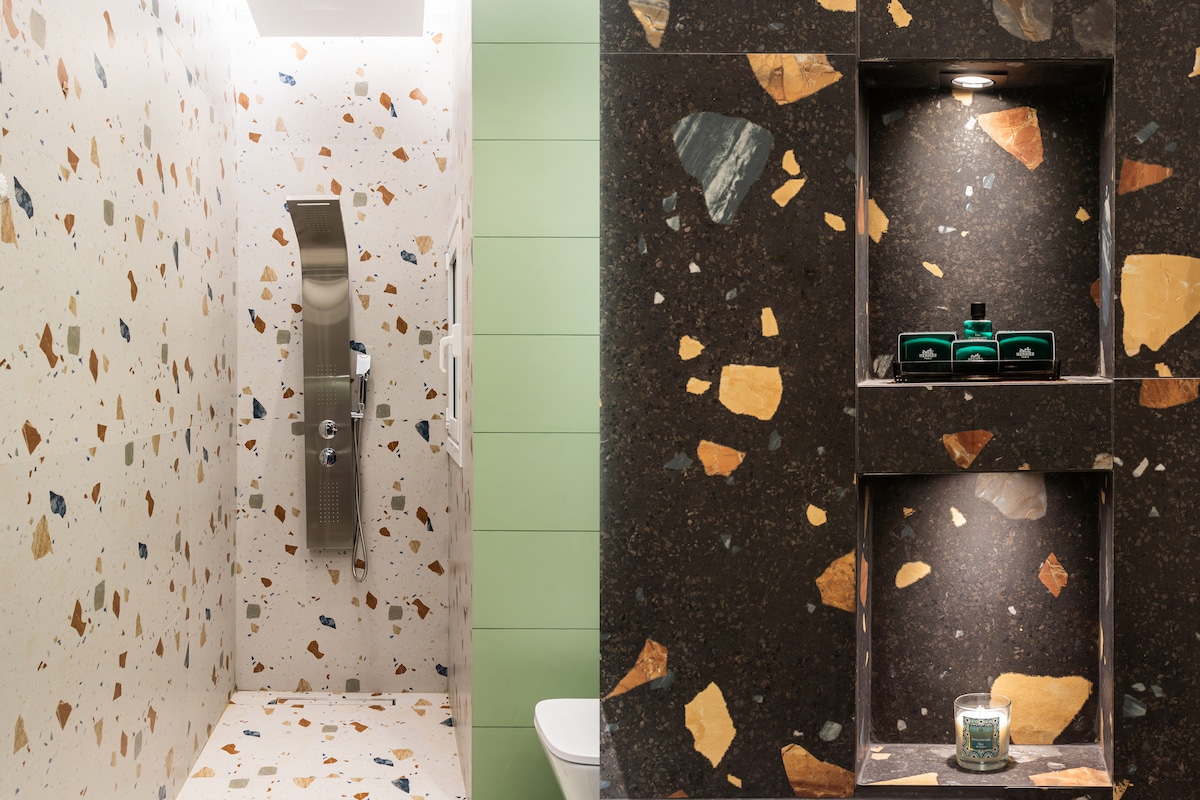 A modern shower area is adorned with a uniquely patterned terrazzo wall, showcasing an array of earthy tones. A sleek shower fixture is installed, complemented by built-in niches that elegantly display toiletries and a decorative candle.