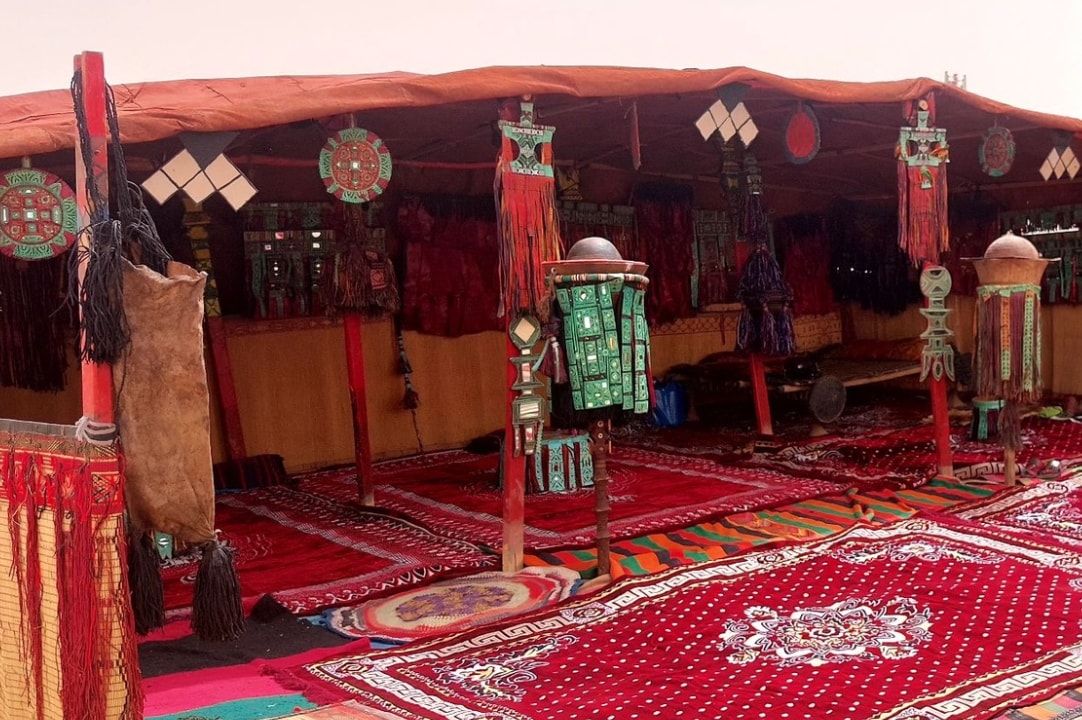 A traditional tent interior showcases colorful textiles and intricate designs. Vibrant rugs cover the ground, while decorative lanterns and patterned wall hangings add visual interest. Wooden poles support the structure, creating an inviting atmosphere for gatherings.