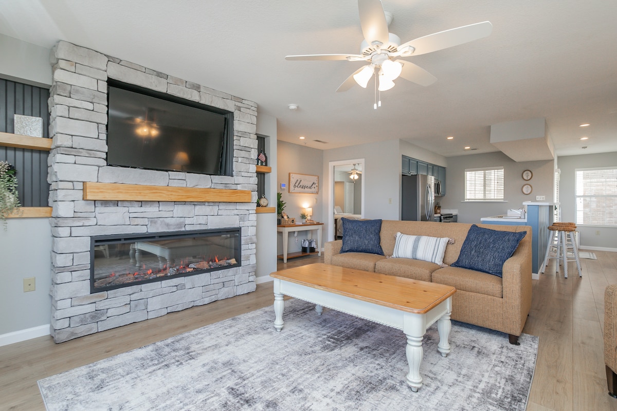 A warm and inviting living area features a stone accent wall with a mounted television above a modern electric fireplace. A comfortable beige sofa is complemented by a rustic wooden coffee table. A patterned area rug anchors the seating space, with a view into the kitchen area.