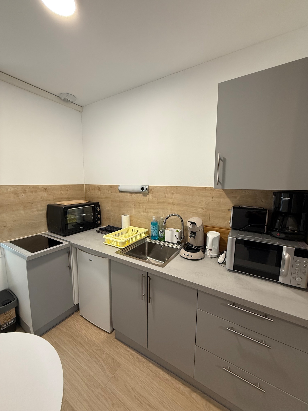 The kitchenette features a modern design with grey cabinetry and a neutral countertop. It includes essential appliances such as a microwave, coffee maker, kettle, and a mini refrigerator. A stainless steel sink is situated between distinct cooking areas, complemented by a dish drying rack.