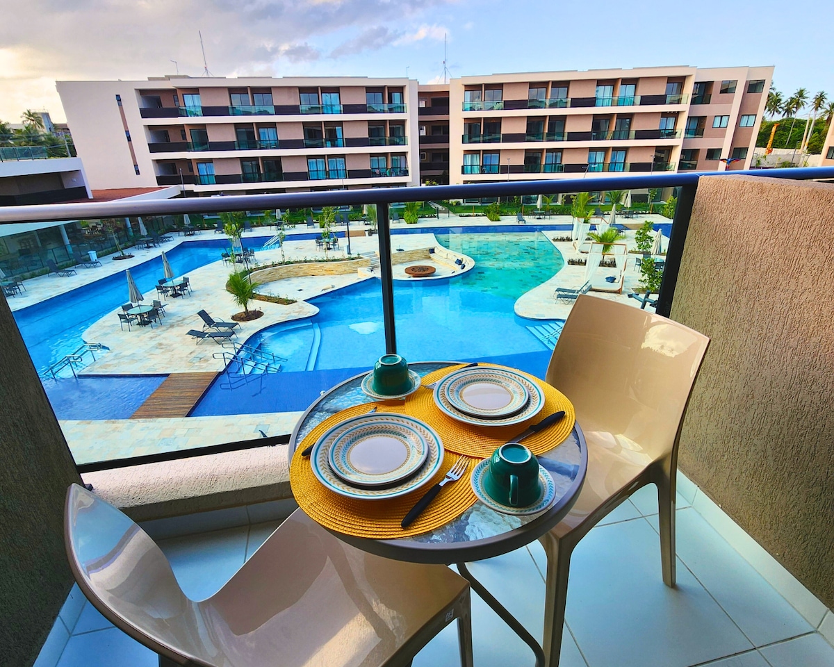 A balcony table is set for two, featuring turquoise cups and plates on a vibrant yellow placemat. The view beyond includes a large pool area, showcasing various swimming pools and lounging spaces surrounded by greenery.