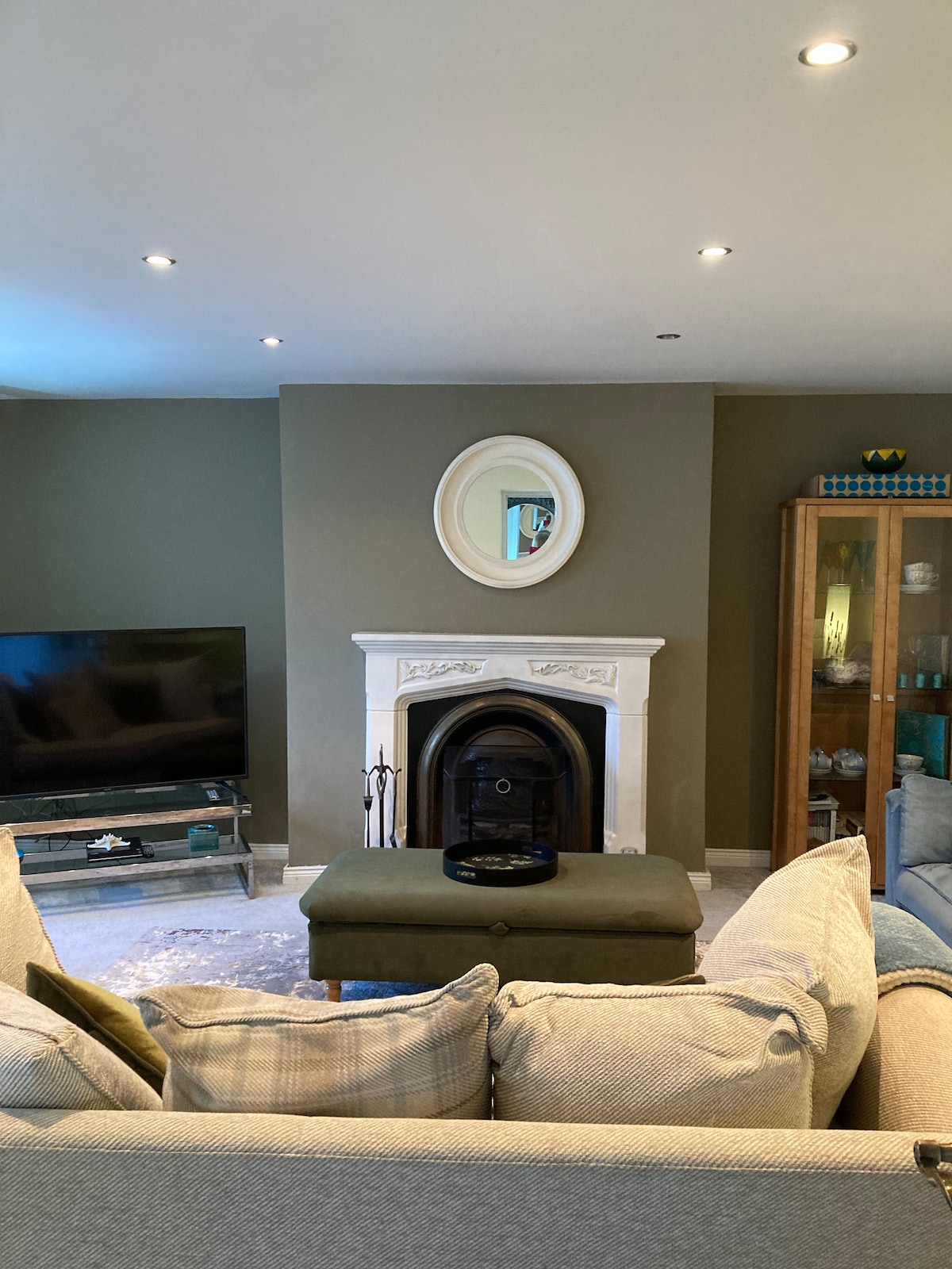 A living area features a minimalist design with a soft grey color palette. A traditional fireplace is framed by an elegant white mantelpiece, while a circular mirror hangs above it. A television rests on a dark media cabinet, and a cozy seating arrangement is visible.