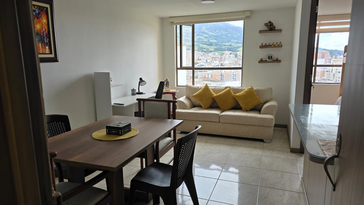 A well-lit living area features a comfortable couch adorned with yellow cushions, a dining table with chairs, and a work desk beside a large window. The view includes distant mountains and cityscape, while tile flooring enhances the spacious feel.