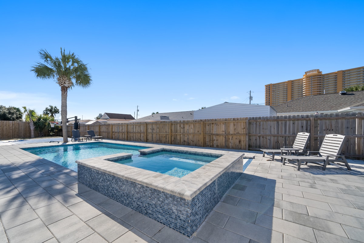 A private backyard features a heated pool surrounded by a stone patio. Lounge chairs are positioned nearby, and palm trees provide a touch of greenery. A wooden fence creates privacy and minimal structures are visible in the background against a clear blue sky.