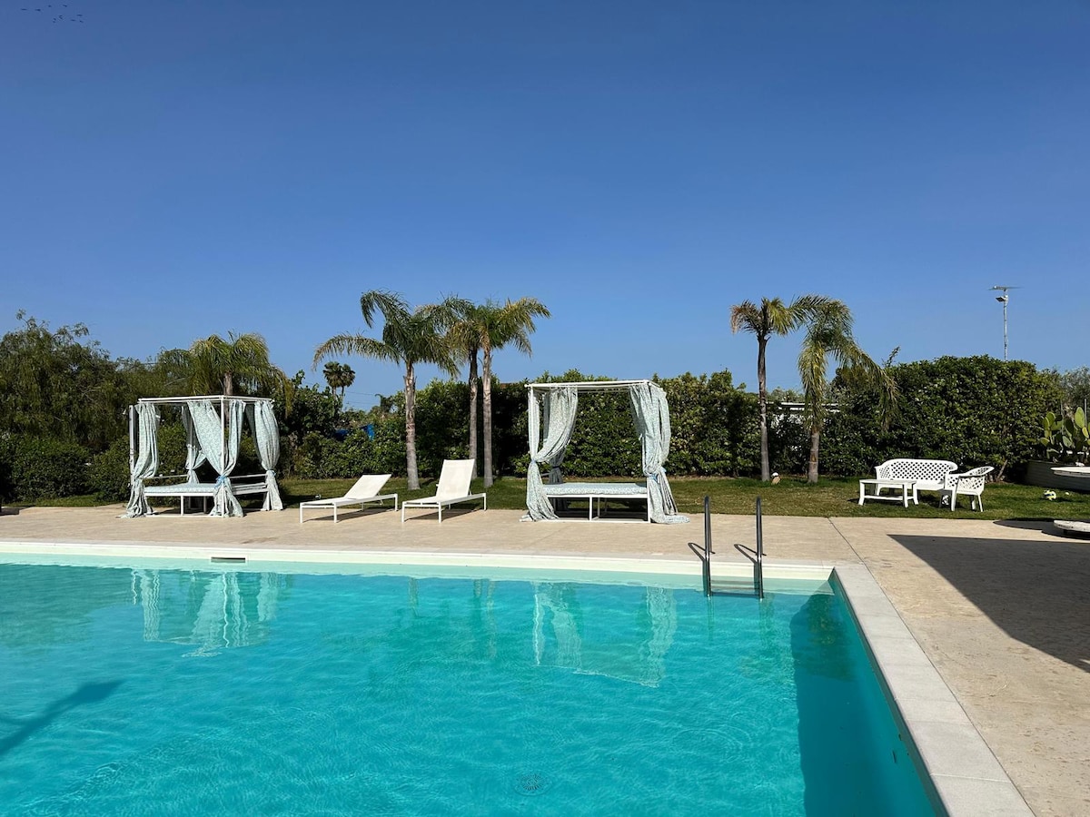 The outdoor area features a refreshing swimming pool surrounded by well-maintained greenery. Comfortable lounge chairs are positioned near the water, while two shaded seating areas with draped fabric offer places to relax and enjoy the sun.