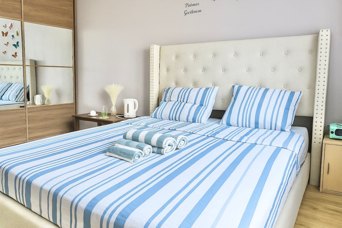 Elegantna spavaća soba s bračnim krevetom i tapeciranim uzglavljem, plavo-bijelom prugastom posteljinom koja daje dojam svježine i modernosti. Savršeno za opuštajući boravak u urednom okruženju.

