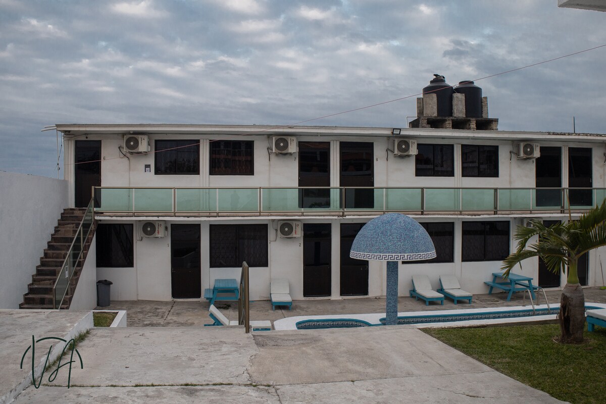 A two-story building features a row of guest rooms with air conditioning units visible. Outdoor lounge chairs are arranged beside a pool, complemented by an umbrella with a blue mosaic design. Access to the upper level is provided by a set of stairs.