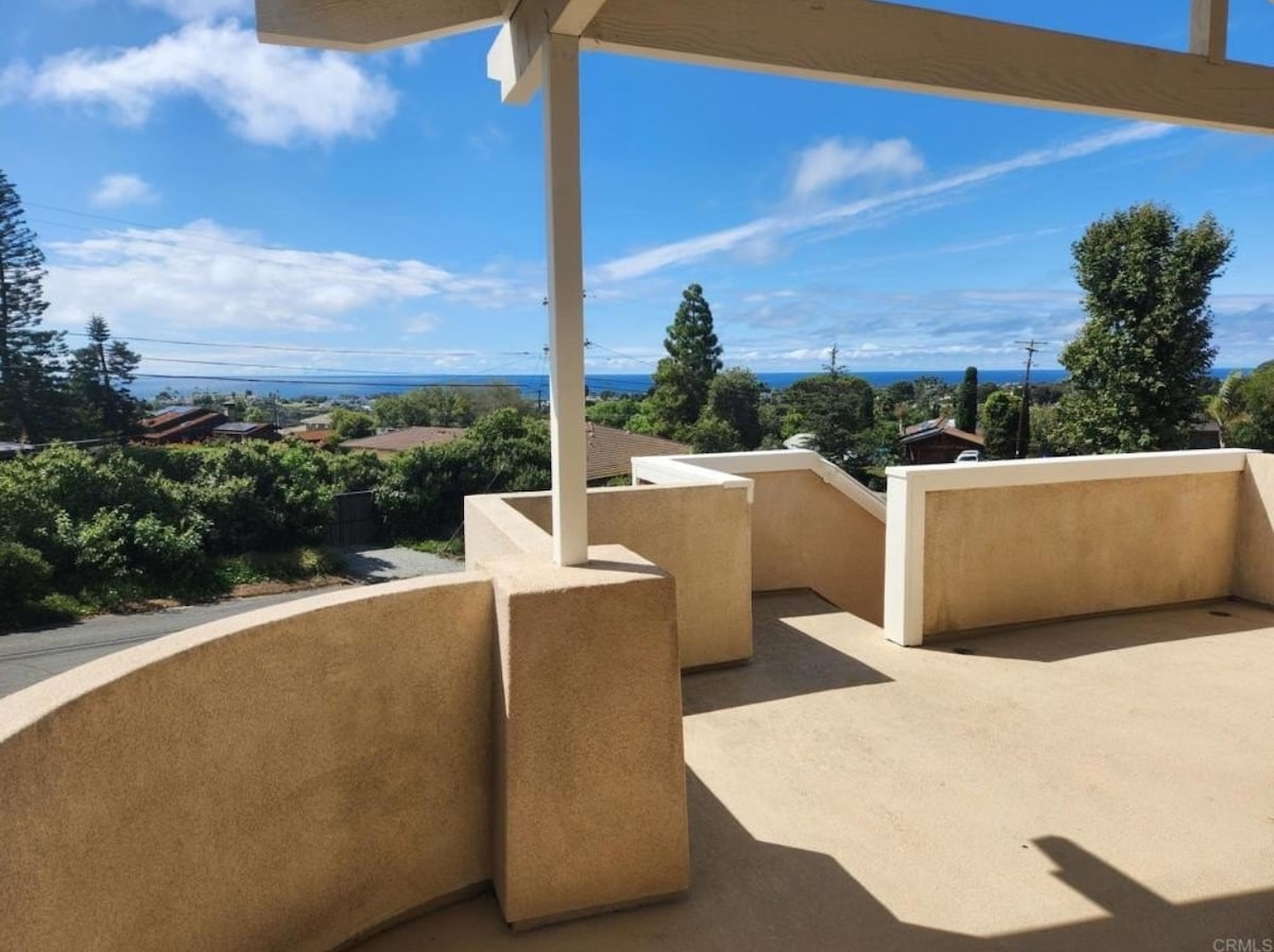 A spacious outdoor balcony is shown, featuring a smooth concrete surface and a protective overhang. Lush greenery and trees frame the view, which opens up to a calming expanse of the ocean and distant horizon under a clear blue sky.