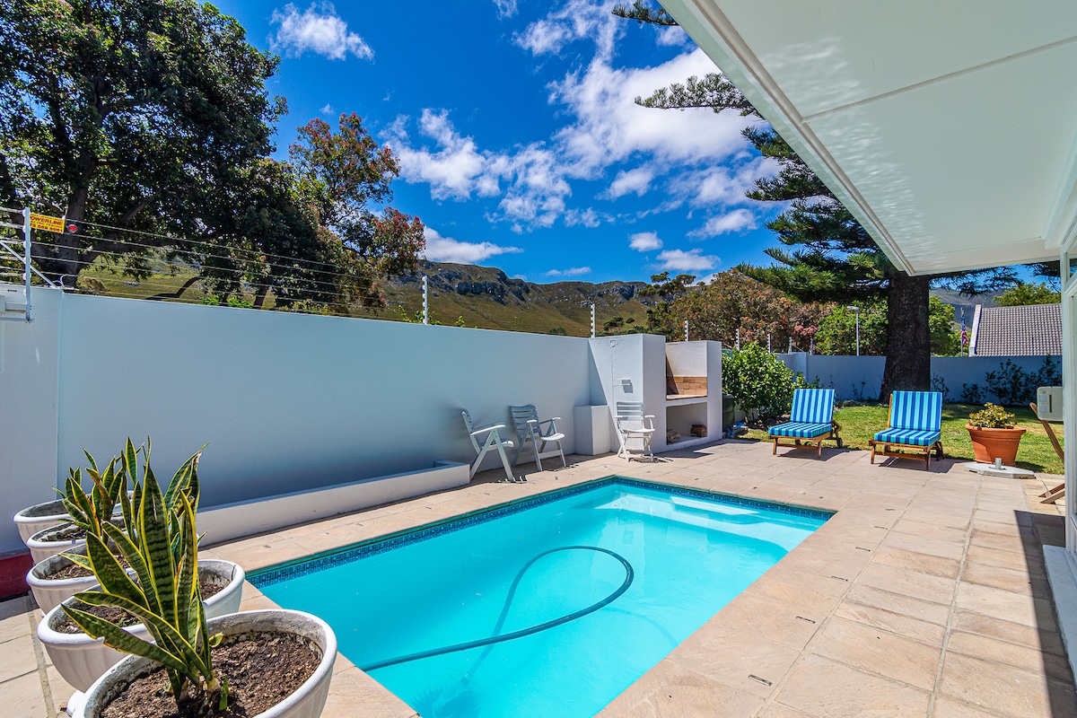 An inviting outdoor area features a swimming pool surrounded by stone tiles. Two lounge chairs are positioned beside the pool, while potted plants add a touch of greenery. The backdrop reveals a clear blue sky and rolling hills in the distance.