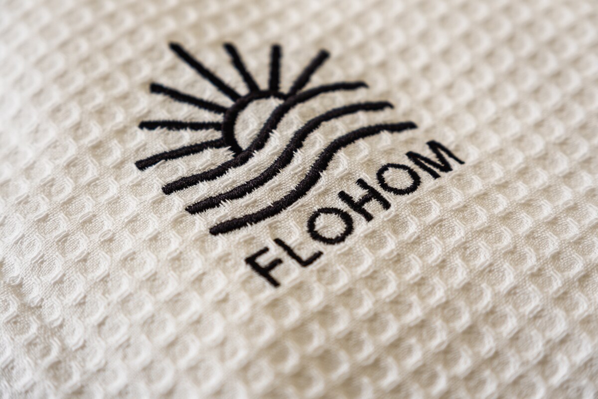 An embroidered logo of FLOHOM is displayed on a textured fabric, featuring a sun and wave design. The logo is presented in dark thread on a soft, light-colored background, emphasizing the brand's connection to water and relaxation.