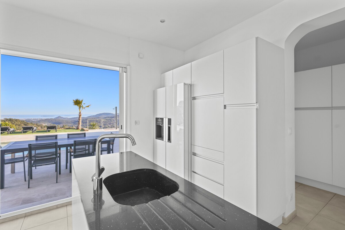 A modern kitchen is showcased with sleek white cabinetry and a dark countertop featuring a deep sink. Large glass doors invite natural light and offer views of an outdoor dining area beyond, framed by a landscape of hills and distant mountains.