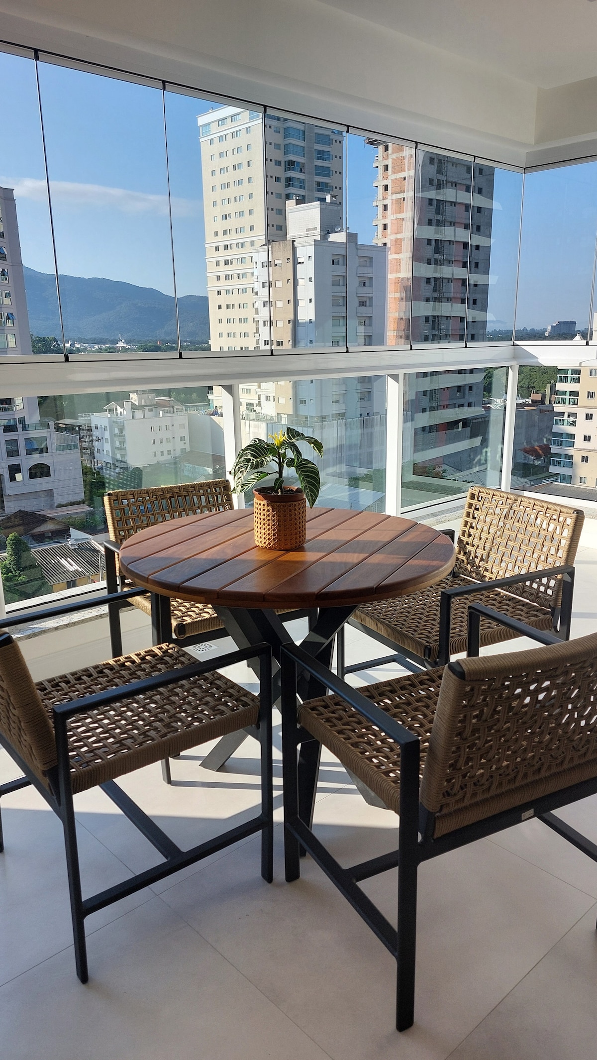 A balcony area is presented with a round wooden table and four woven chairs. A small potted plant is placed at the center of the table, surrounded by clear glass panels that offer views of the buildings and mountains in the distance.
