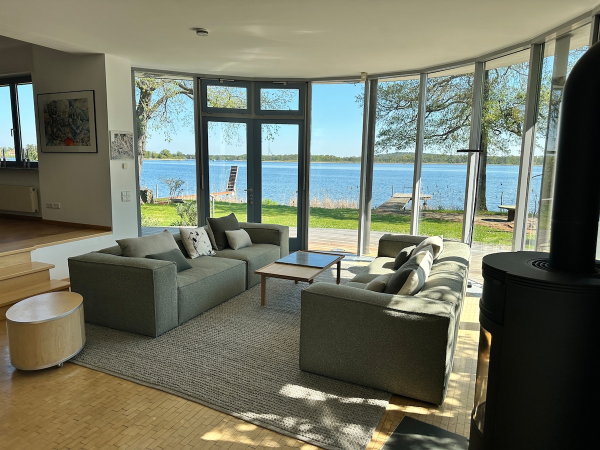 A bright living area features large glass windows offering views of the water and surrounding nature. Comfortable seating is arranged around a central coffee table, and a cozy area rug adds warmth to the space. A modern wood stove is positioned nearby.