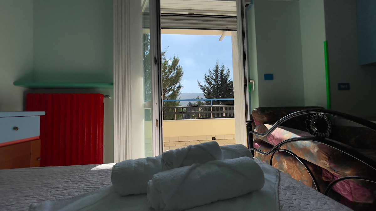 A comfortable room features a spacious bed with neatly rolled towels placed on top. Large glass doors open to a balcony, offering views of greenery outside. A colorful sofa and a light blue cabinet are visible, contributing to a relaxed atmosphere.