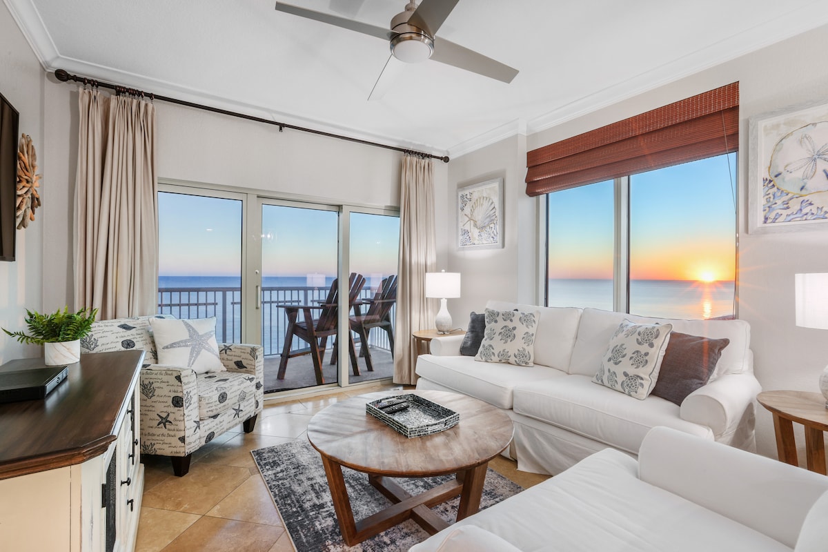 The living area features a spacious white sofa complemented by two patterned chairs. A circular wooden coffee table sits in the center, and large windows reveal a stunning view of the sunset over the Gulf. Light curtains frame the windows, enhancing the serene atmosphere.