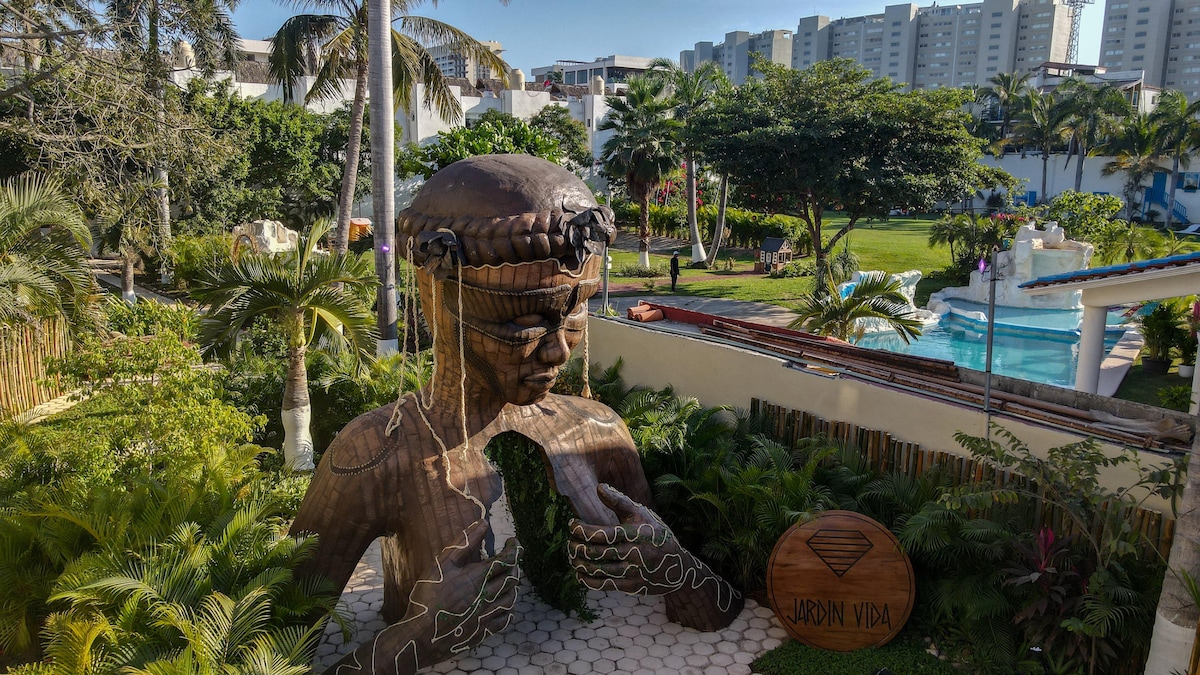 A wooden sculpture of a serene figure is featured in a lush garden setting, surrounded by palm trees and vibrant greenery. The background includes a swimming pool and modern buildings, creating a blend of nature and urbanity.