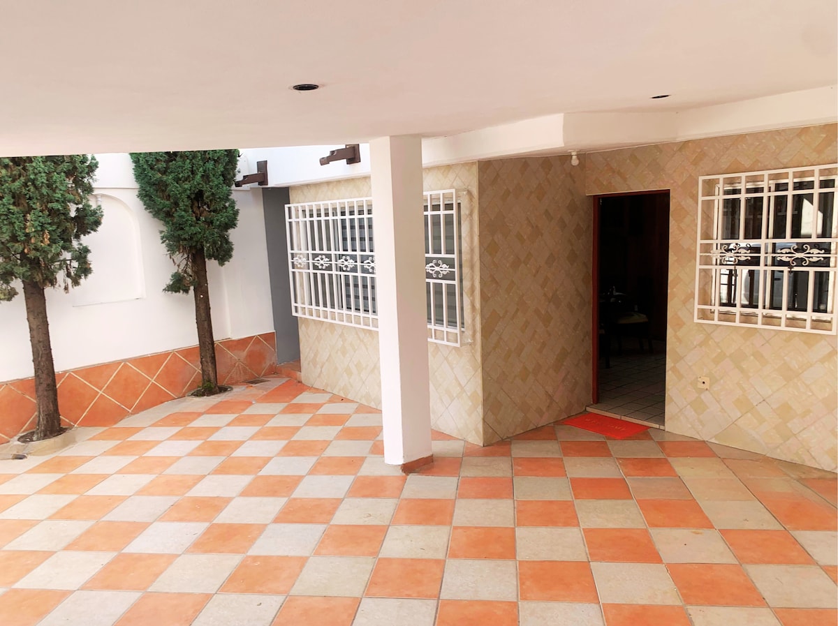 An open courtyard area features a checkered tiled floor in warm hues. Two trees provide greenery against the wall, while a nearby entrance is framed by white bars. Soft natural light enhances the spacious feel of the outdoor space.