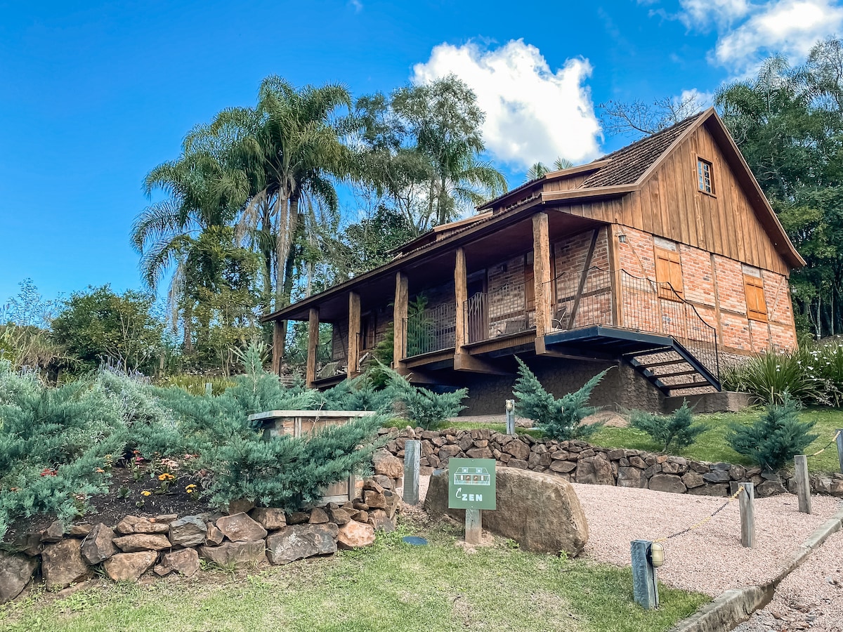 A wooden chalet with a sloped roof is set amidst lush greenery. The structure is elevated on stilts and features a large balcony supported by sturdy columns. Surrounding gardens showcase native plants and rocky landscaping, enhancing the natural atmosphere.