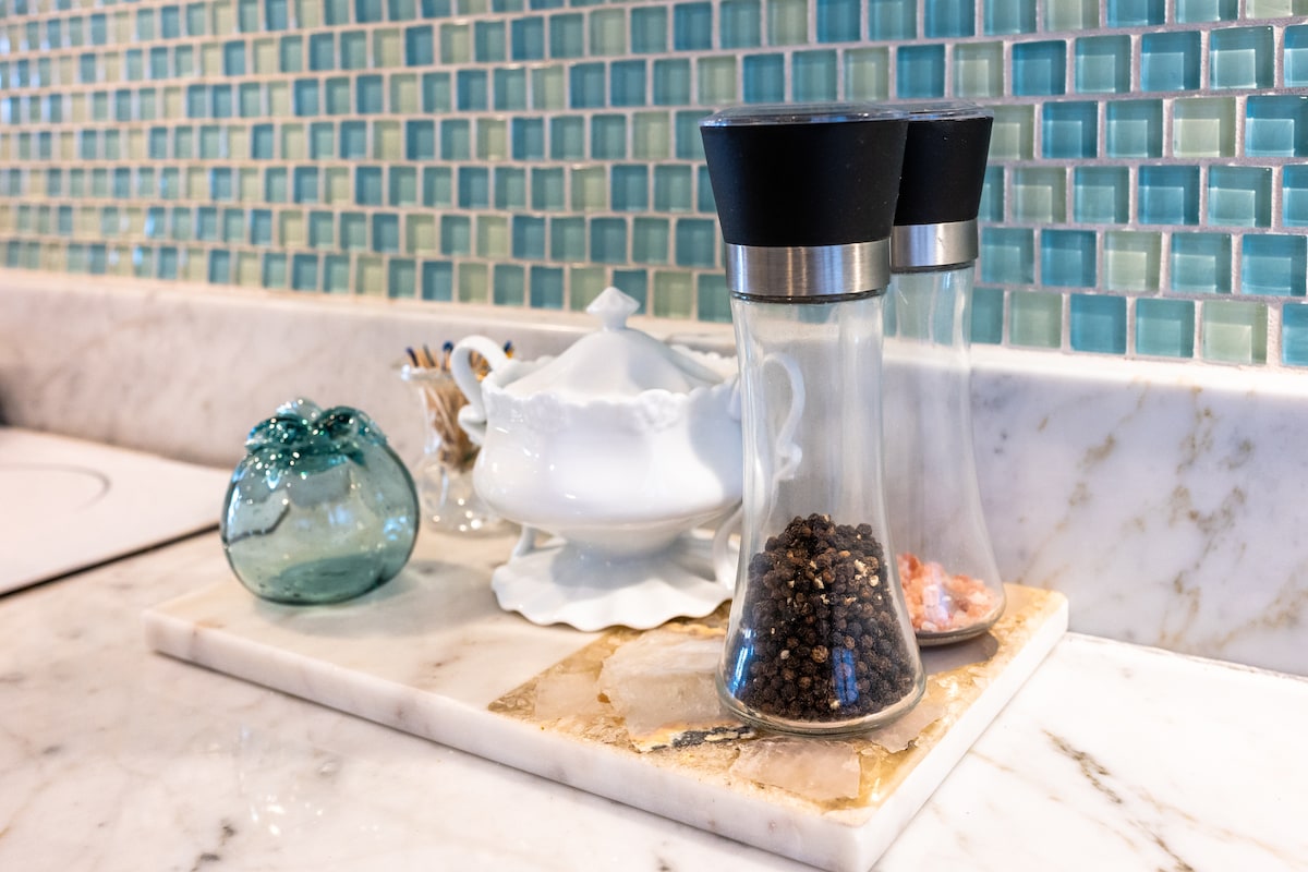 A modern kitchen counter displays two glass spice containers, one filled with black pepper and another with pink salt. A delicate white serving dish is situated nearby, alongside an elegant glass decorative piece in a soft blue hue. The backdrop features a textured aqua tile wall.
