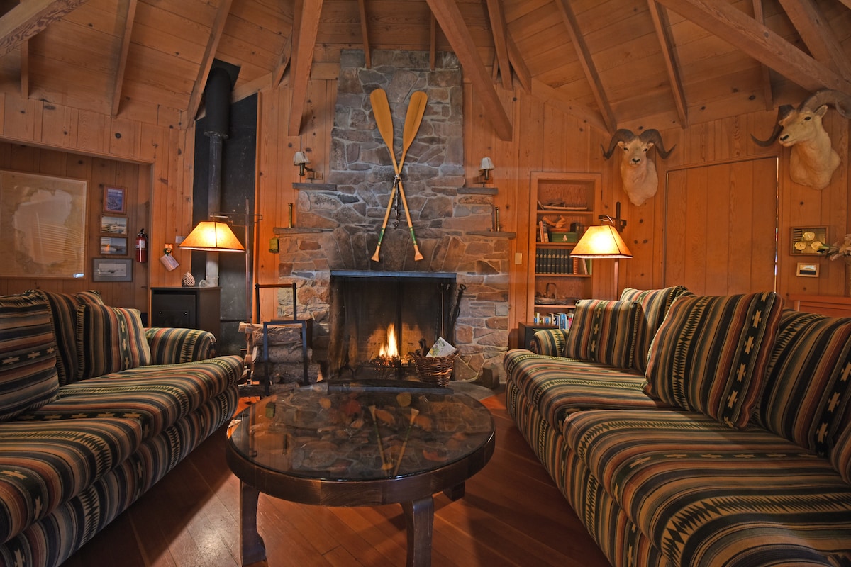 The inviting living room features a stone fireplace as the focal point, flanked by decorative paddles. Two striped sofas face each other around a round coffee table with a stone base. Warm lighting from table lamps enhances the cabin's rustic charm, highlighted by wooden beams and paneling.