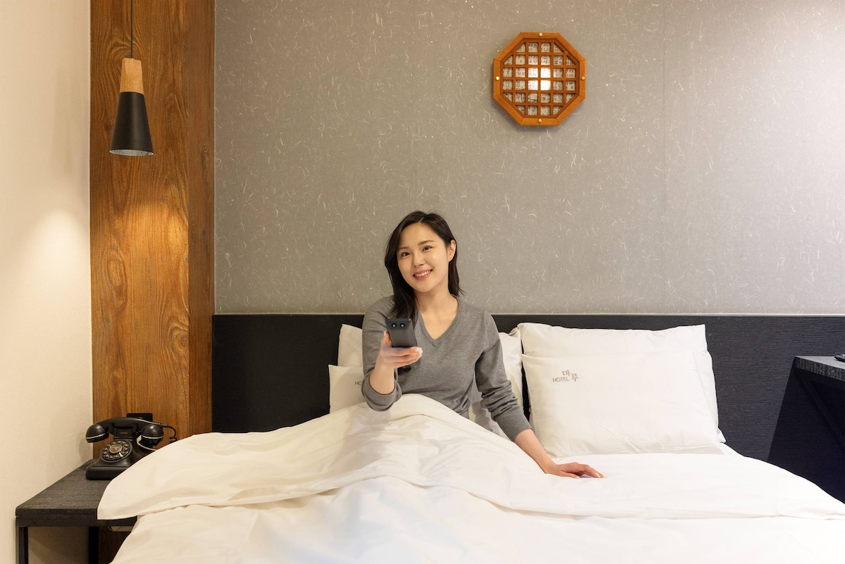 A spacious bed is covered with a neatly arranged white duvet, complemented by decorative pillows. A person seated on the bed holds a remote. Soft lighting emanates from a wall-mounted lamp, while a textured wall and a wooden accent complete the serene atmosphere.