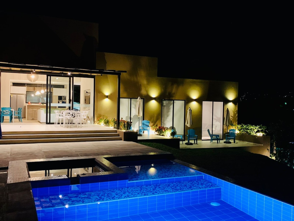A spacious outdoor area is featured, illuminated by soft lighting, highlighting the inviting swimming pool. A jacuzzi is adjacent to the pool, with lounge chairs positioned around. The entrance to the home reveals large glass doors leading into an open dining and kitchen space.