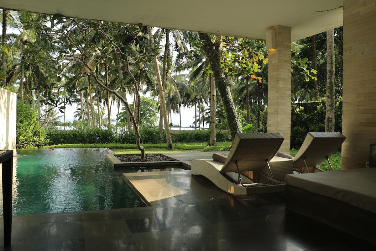 A serene outdoor space features two sun loungers beside a tranquil pool, surrounded by lush greenery and palm trees. The view extends to distant ocean waves, framed by a modern architectural structure with natural stone elements.