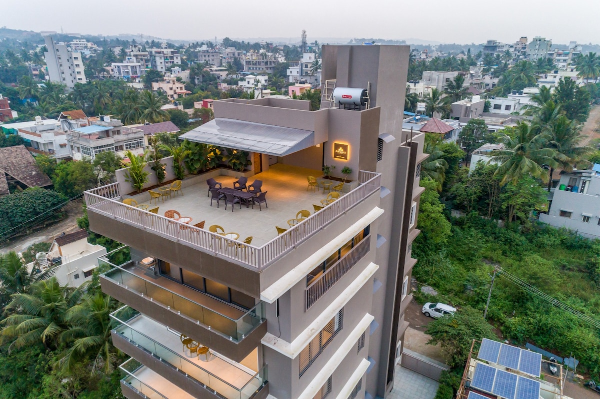 The exterior view showcases a modern multi-floor building featuring a spacious balcony adorned with seating arrangements. Lush greenery surrounds the property, with city rooftops visible in the background. The clean lines and contemporary architecture highlight the luxury accommodations nearby.