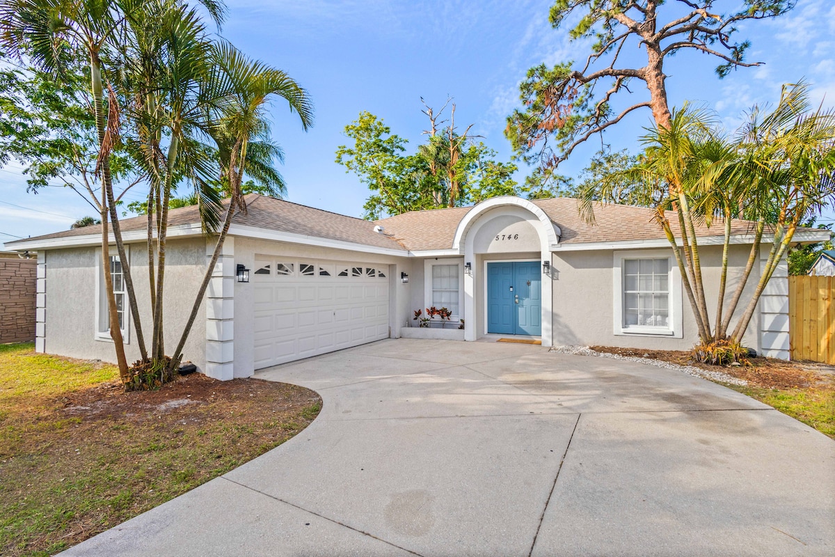A single-story house is presented, featuring a light-colored exterior and a charming blue front door. A spacious driveway leads up to the garage. Palm trees and other greenery frame the entrance, contributing to a welcoming curb appeal.