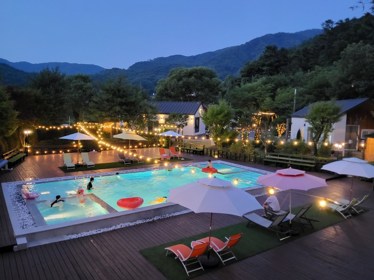 An inviting outdoor pool area is illuminated with warm lights, surrounded by lush greenery and mountains. Umbrellas provide shade over lounge chairs, while guests enjoy the refreshing water. Colorful floats and a wooden deck enhance the relaxing atmosphere of the inviting space.