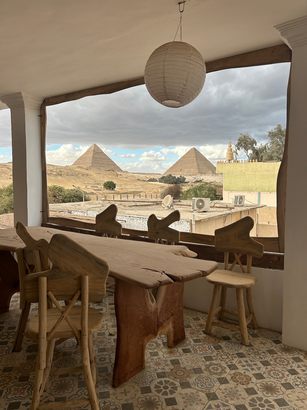 A rustic dining area features a large wooden table with natural finishes, accompanied by uniquely shaped wooden chairs. Large windows provide expansive views of the pyramids in the distance, while a pendant light hangs overhead, illuminating the space.