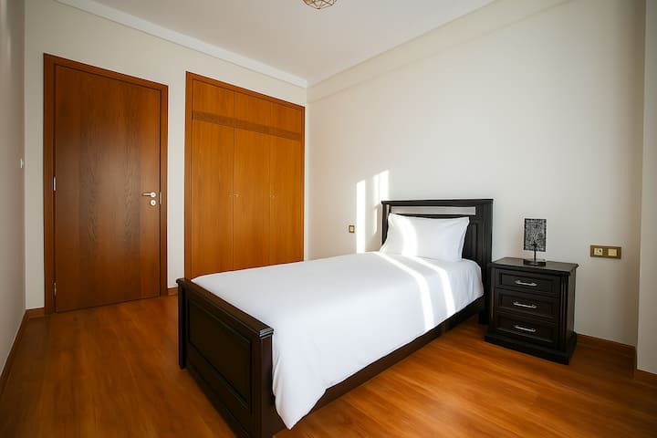 Un cómodo dormitorio con una cama individual con colchón adicional, mucha luz y persianas enrollables automáticas. Sube al balcón para disfrutar de la vista del parque verde y relájate con el desayuno en la mesa y las sillas.