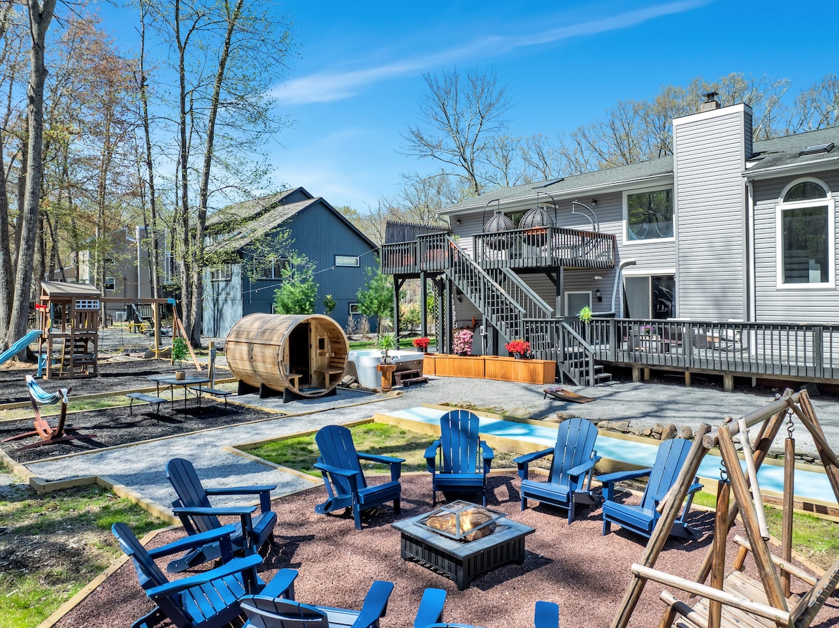 An outdoor space is showcased, featuring a fire pit surrounded by six blue chairs. A barrel sauna is positioned nearby, with a playground visible in the background. The two-story house and deck provide a spacious backdrop against a clear blue sky and wooded area.