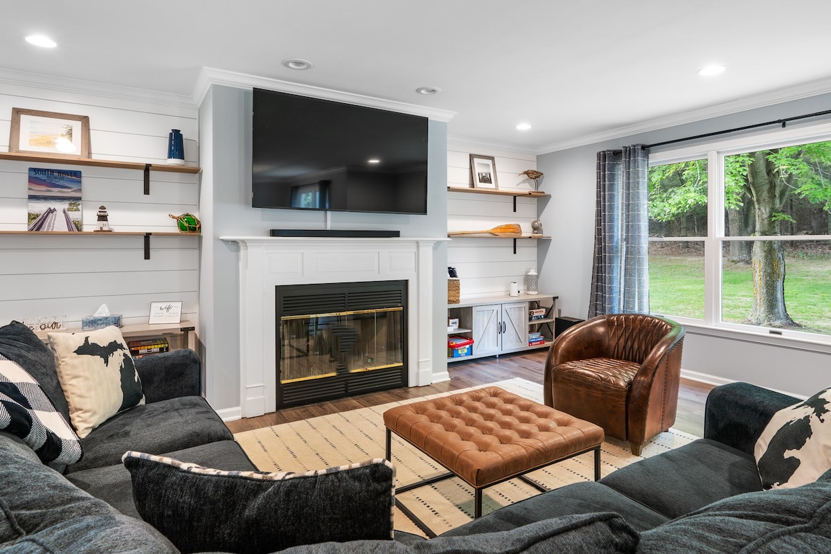 A cozy living room features a sectional sofa, a leather armchair, and a coffee table. A large TV is mounted above a modern fireplace. Natural light enters through the window, offering a view of the backyard, while shelves display decorative items and framed photographs.