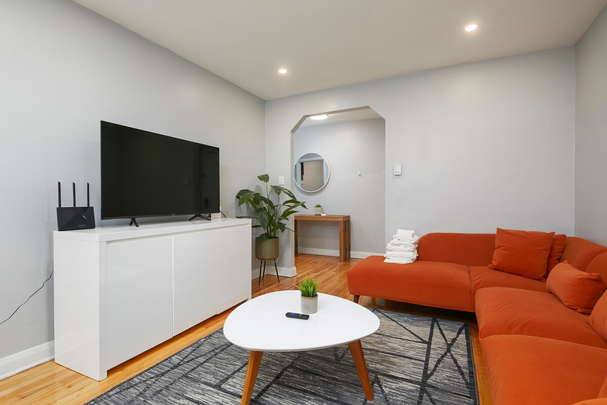 A modern living area features a vibrant orange sofa positioned around a white coffee table. A large television rests atop a white cabinet. Natural light fills the space, complemented by a green plant and a circular mirror on the wall.
