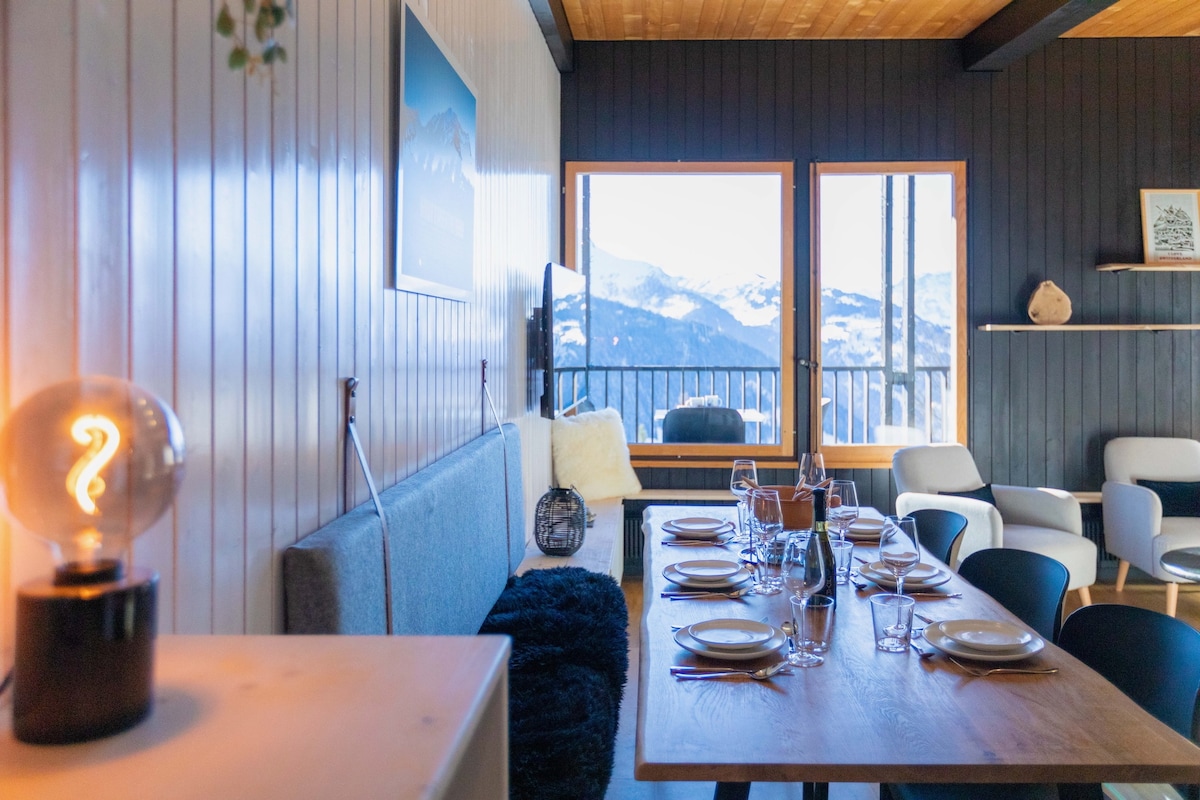 A dining area features a long wooden table set with plates and cutlery, surrounded by black chairs. Large windows provide views of the mountains, while a soft grey sofa, illuminated by a unique lamp, creates a welcoming space. Neutral tones enhance the modern alpine design.