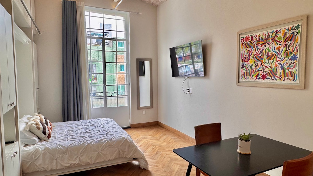 A bright and functional loft space is depicted, featuring a spacious bed with a textured white coverlet. A wooden dining table with two chairs is positioned near a flat-screen TV mounted on the wall. Large windows allow natural light to fill the room, complemented by light-colored curtains.