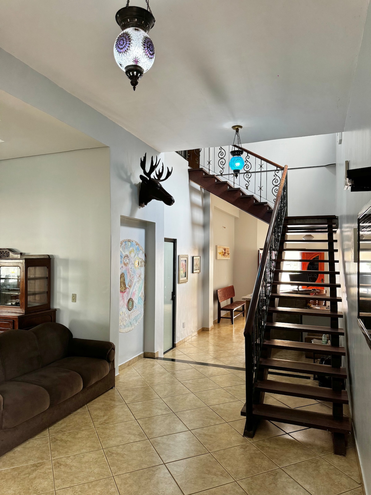 A well-lit entryway features a spiral staircase leading to an upper floor. Decorative lanterns hang from the ceiling, and a mounted animal head adds character to the wall. A cozy sofa and a wooden chair are positioned for seating along the tiled floor.
