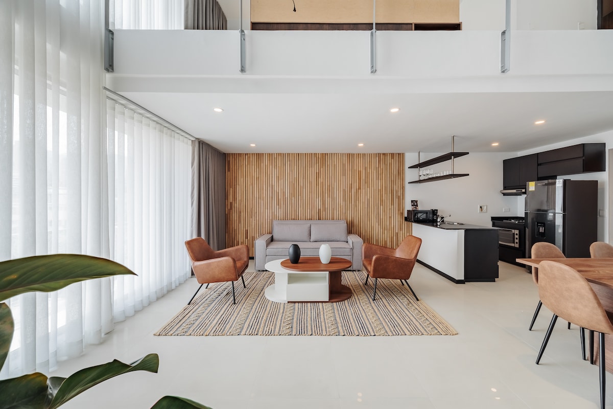 A contemporary living area features a light gray sofa facing a round coffee table placed on a textured rug. Two brown accent chairs are positioned nearby. The space is illuminated by natural light filtering through sheer drapes, creating a warm ambiance against a backdrop of wooden wall paneling.