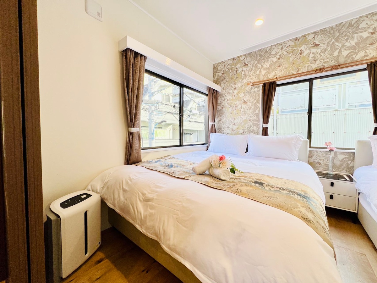 A private guest room features two double beds adorned with neutral-toned bedding. Large windows allow natural light to fill the space, framed by light curtains. A small air purifier is positioned beside the bed, contributing to a comfortable atmosphere.