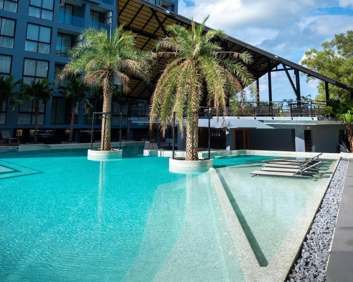 A spacious pool area features a tranquil, turquoise water surface bordered by elegant palm trees. Lounge chairs are positioned on the poolside, and a stylish structure with a roof frames the backdrop, enhancing the serene environment.