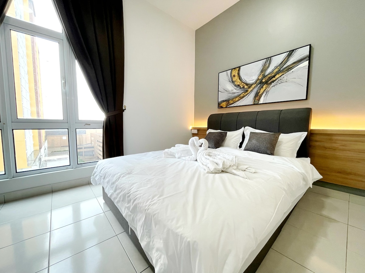 A spacious bedroom features a large bed dressed in fresh white linens, with decorative pillows arranged on top. Light filters through the floor-to-ceiling window, where dark curtains are elegantly drawn to the side. A piece of abstract art adds a modern touch to the wall.