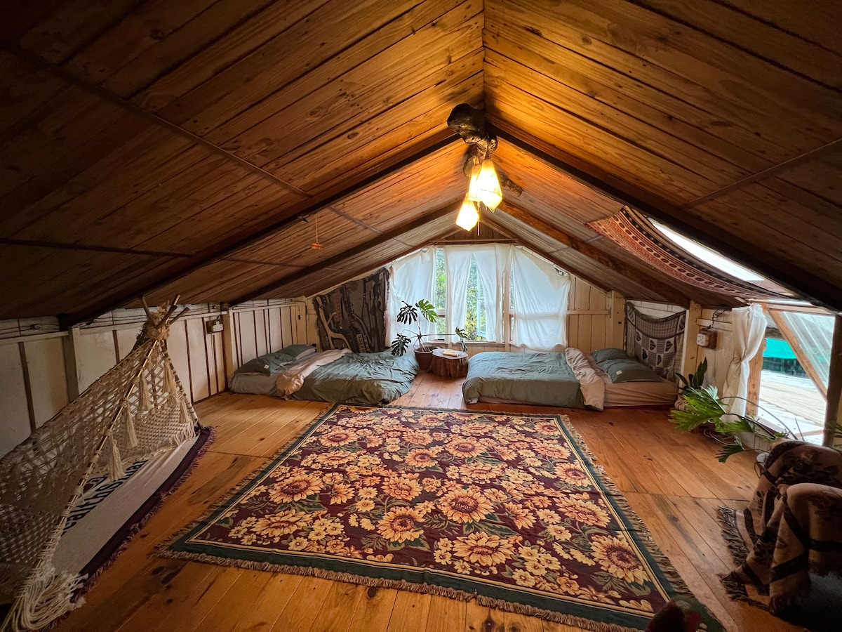An attic room with wooden walls and ceiling features two queen mattresses arranged on a wooden floor. A colorful area rug sits in the center, while natural light filters through large windows. A hammock is visible in one corner, adding to the relaxed atmosphere.