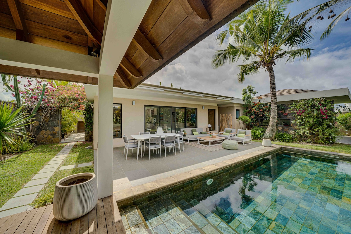 An outdoor terrace features a rectangular pool surrounded by lush greenery and tropical plants. A spacious dining table is paired with several lounge chairs, providing ample seating for relaxation. The patio is shaded by a wooden overhang, offering a comfortable space for enjoying the outdoors.