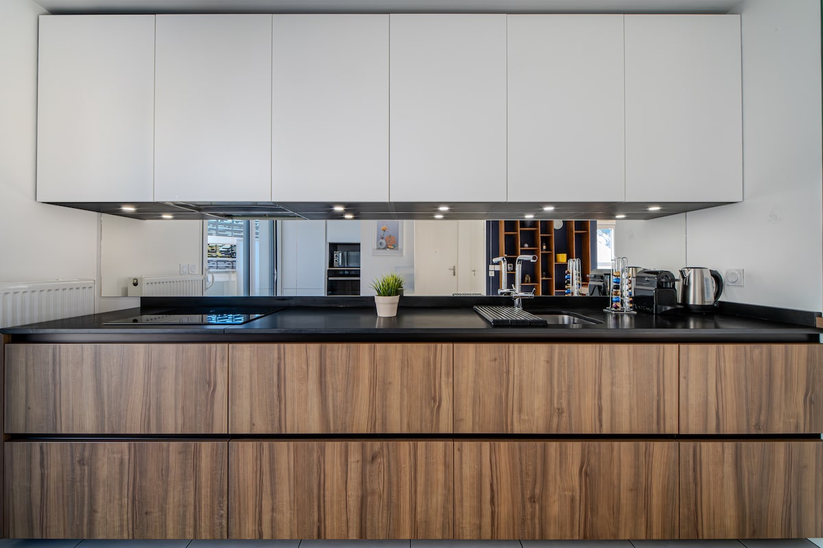 A modern kitchen features sleek cabinetry with a combination of white upper units and warm wood lower drawers. Counter space is highlighted by a black countertop, and a small plant adds a touch of greenery. Appliances include a kettle and a built-in cooktop.
